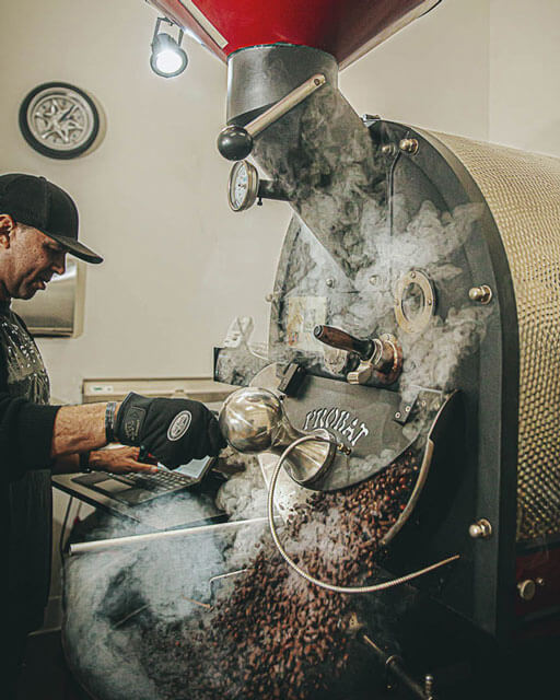 Black Stallion Coffee owner completing the whole bean coffee roasting process from his Midnight Rendezvous coffee blend.  Aromatizing coffee bean steam is bursting out of coffee roasting machine filling the air with a black cherry, toasted hazelnut aroma 